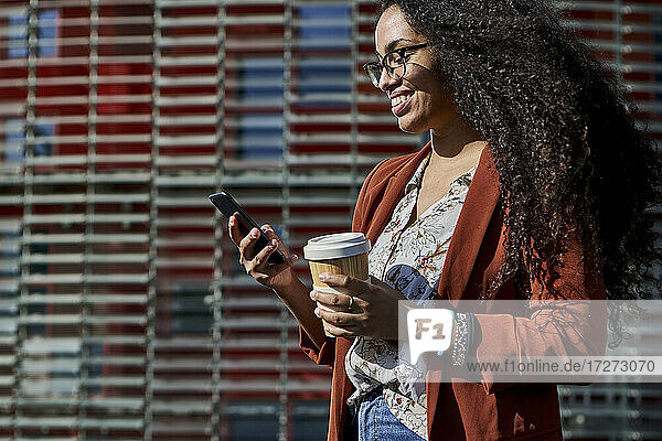 Lächelnde Frau  die ein Smartphone benutzt und einen wiederverwendbaren Becher hält  während sie vor einem Gebäude steht