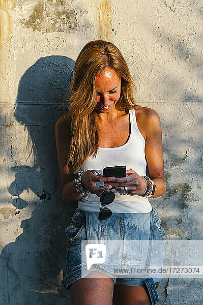 Lächelnde Frau  die ein Mobiltelefon benutzt  während sie an der Wand steht