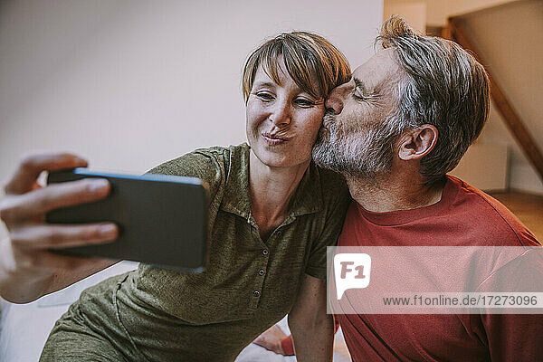 Reifer Mann küsst Frau  während er ein Selfie auf dem Smartphone macht  das im Schlafzimmer zu Hause steht