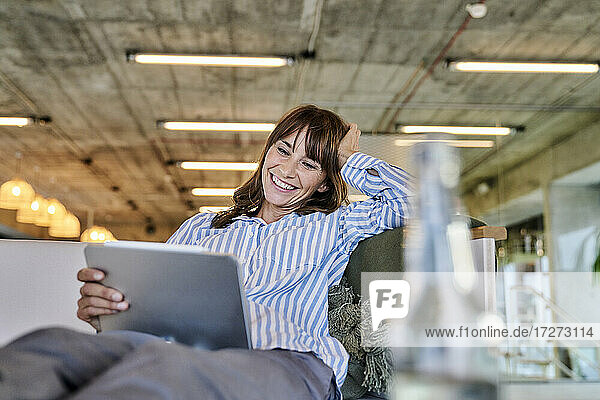 Lächelnde Frau mit Hand in Händen  die ein digitales Tablet benutzt  während sie zu Hause auf dem Sofa sitzt