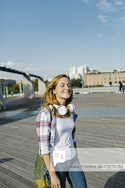Glückliche Frau mit Kopfhörern um den Hals  die an einem sonnigen Tag auf einem Fußweg spazieren geht