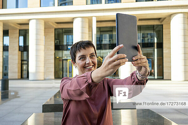 Lächelnde Frau  die ein Selfie mit einem digitalen Tablet macht  während sie an einem Gebäude in der Stadt sitzt