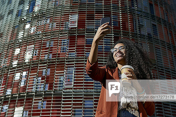 Lächelnde junge Frau führt einen Videoanruf mit ihrem Smartphone durch  während sie an einem sonnigen Tag vor einem Gebäude steht