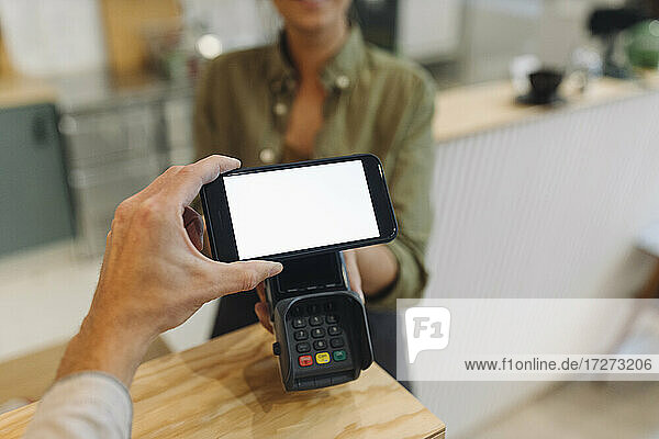 Abgeschnittene Hand eines Kunden  der mit seinem Smartphone kontaktlos bezahlt  während die Besitzerin ein Kreditkartenlesegerät an der Kasse eines Cafés hält
