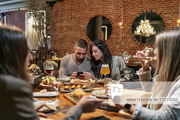 Women talking while drinking wine with friends using mobile phone in background at restaurant