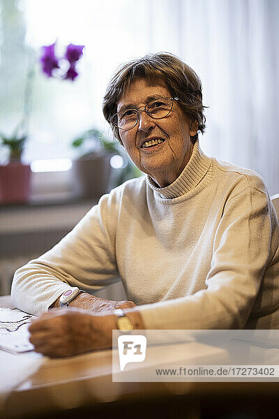 Smiling wrinkled woman with emergency button on wrist sitting by table at home