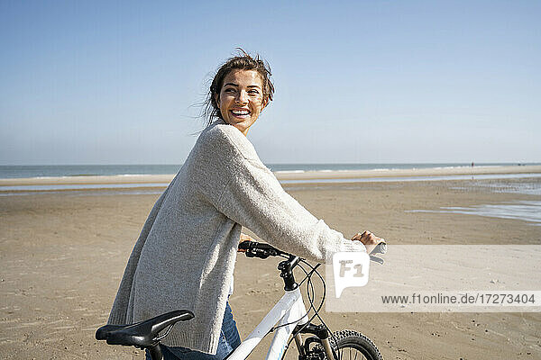 Fröhliche junge Frau  die mit dem Fahrrad spazieren geht und über die Schulter auf den Strand schaut  an einem sonnigen Tag