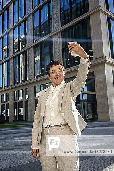 Lächelnde Frau nimmt Selfie im Stehen gegen moderne Bürogebäude