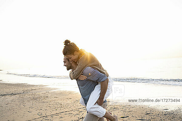 Lächelnder Mann  der seine Freundin huckepack nimmt  während er am Strand gegen den klaren Himmel bei Sonnenuntergang spazieren geht