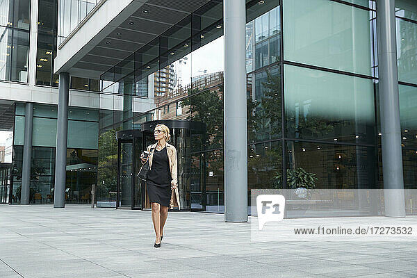 Businesswoman holding coffee cup while walking against office building