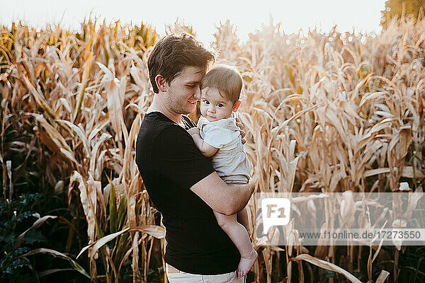 Father carrying baby boy while standing against field during sunset
