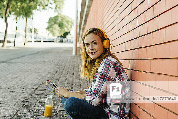 Frau hört Musik auf dem Smartphone über Kopfhörer  während sie an einer Mauer sitzt