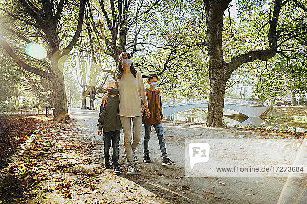 Mutter und Söhne tragen einen Gesichtsschutz beim Spaziergang in einem öffentlichen Park an einem sonnigen Tag