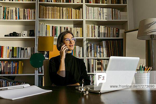 Geschäftsfrau  die mit einem Smartphone spricht und wegschaut  während sie zu Hause am Laptop sitzt und auf ein Bücherregal schaut