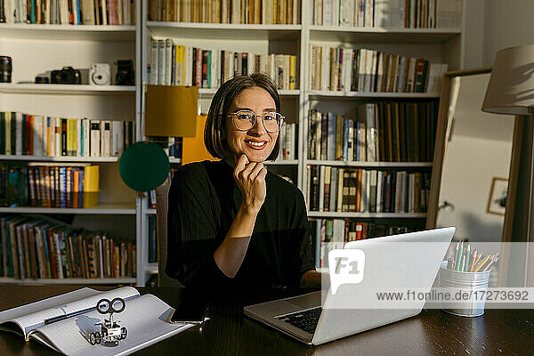 Geschäftsfrau mit Hand am Kinn  die einen Laptop benutzt  während sie an einem Bücherregal sitzt