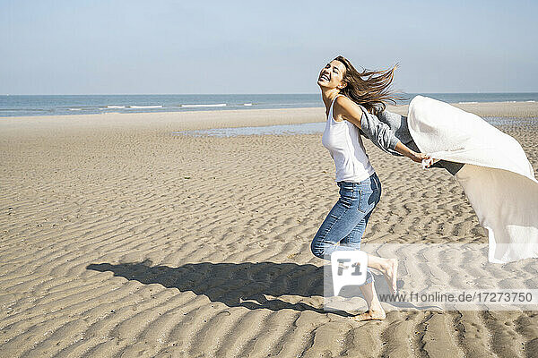 Unbeschwerte junge Frau  die an einem sonnigen Tag am Strand läuft und eine Decke hält