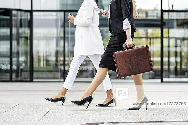 Businesswoman with briefcase using mobile phone while walking by colleague on footpath