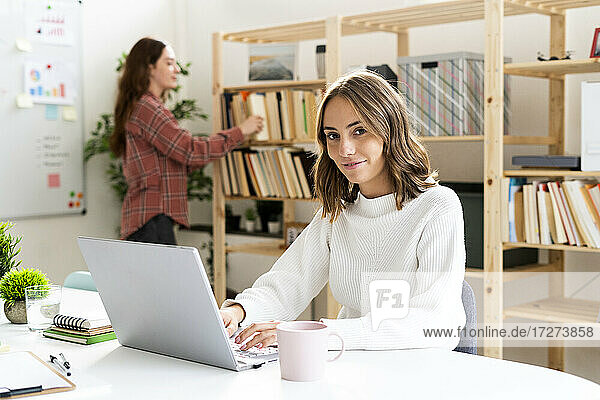 Junge Geschäftsfrau  die einen Laptop benutzt  während ein Kollege im Hintergrund im Büro steht