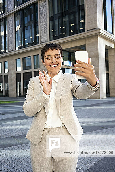 Frau winkt mit der Hand zu einem Videoanruf auf einem Mobiltelefon  während sie vor einem modernen Bürogebäude steht