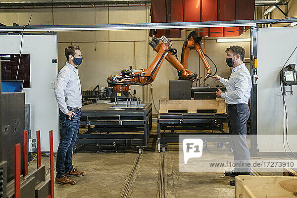 Geschäftsleute mit Gesichtsmaske stehen mit sozialem Abstand vor einer Robotermaschine in einer Fabrik