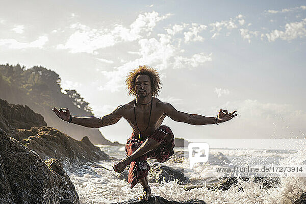 Hemdloser junger Mann übt Entspannungsübungen am Strand gegen den Himmel