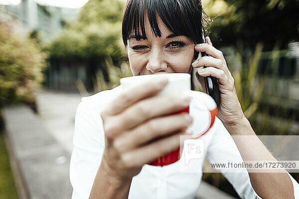 Lächelnde Geschäftsfrau,  die eine Kaffeetasse hält,  während sie mit einem Mobiltelefon spricht