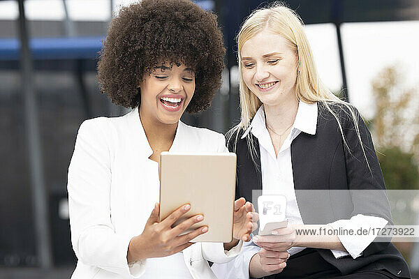 Geschäftsfrauen  die ein digitales Tablet und ein Smartphone benutzen  während sie an einem Gebäude sitzen