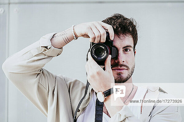 Confident man taking photograph through camera while standing against wall