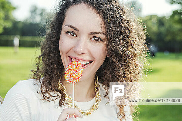 Schöne glückliche junge Frau mit lockigem braunem Haar isst Lutscher im Park