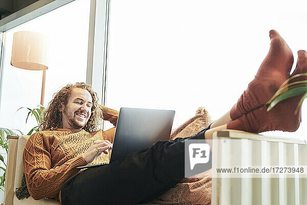Lächelnder Mann  der einen Laptop benutzt  während er zu Hause auf dem Sofa liegt