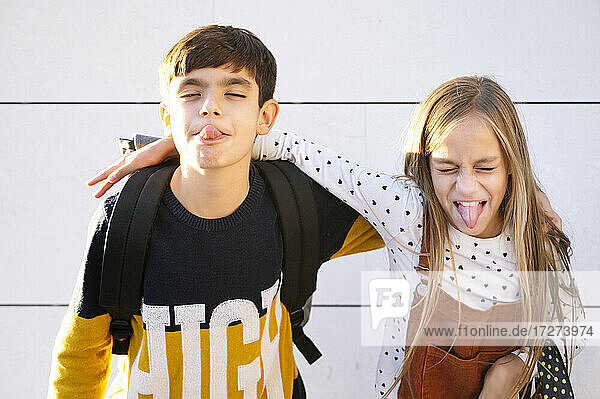 Brother and sister sticking out tongue while standing with arm around against wall on sunny day