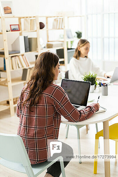 Junge Frau  die einen Laptop benutzt  mit einer Geschäftsfrau  die im Hintergrund im Büro sitzt