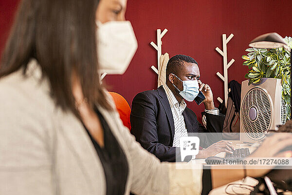 Young businessman in protective face mask talking on mobile phone at office during COVID-19