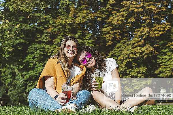 Smiling female friends sitting with fresh lemonade on grass at park during sunny day