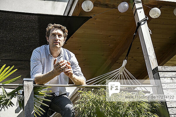 Mann schaut weg  während er mit einer Kaffeetasse auf einem Balkon an einem sonnigen Tag steht