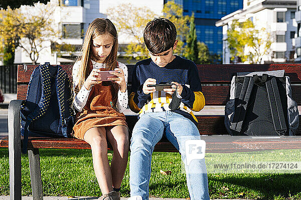 Bruder und Schwester benutzen ihr Smartphone  während sie auf einer Bank in einem öffentlichen Park bei Sonnenschein sitzen