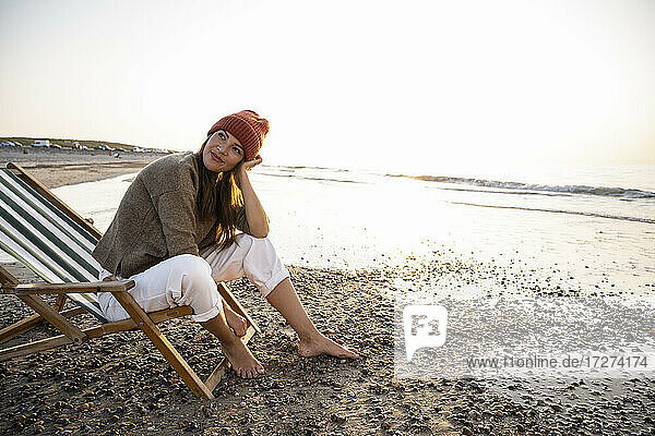 Nachdenkliche Frau  die auf einem Klappstuhl sitzt und bei Sonnenuntergang gegen den klaren Himmel auf den Strand blickt