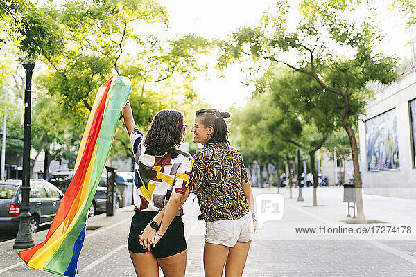 Lesbian couple holding rainbow flag while standing on footpath in city