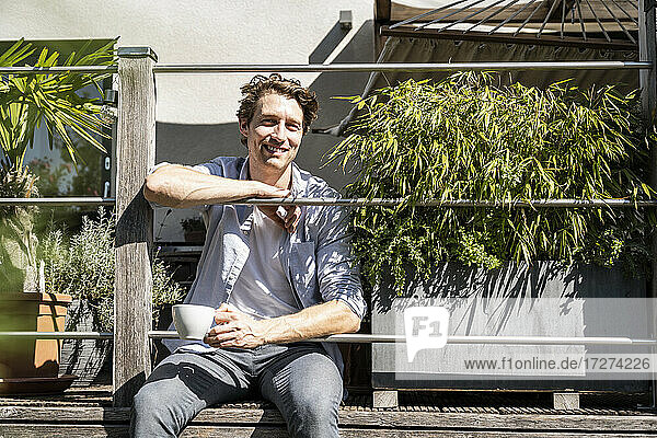 Lächelnder Mann mit Kaffeetasse auf dem Balkon an einem sonnigen Tag