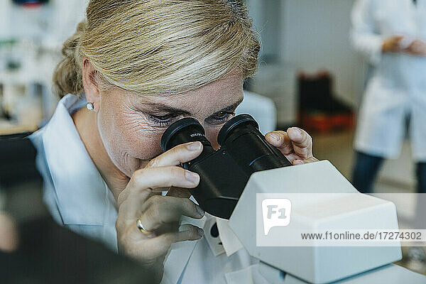Female scientist looking through microscope while man standing in background at laboratory