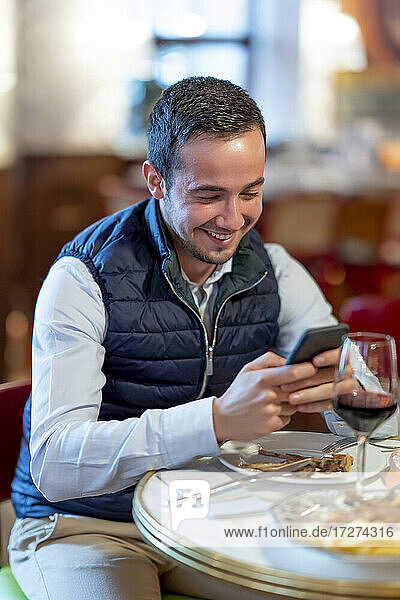 Glücklicher junger Mann  der am Esstisch eines Restaurants eine Textnachricht auf seinem Smartphone schreibt