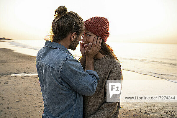 Romantischer junger Mann  der das Gesicht seiner Freundin berührt  während er bei Sonnenuntergang am Strand steht