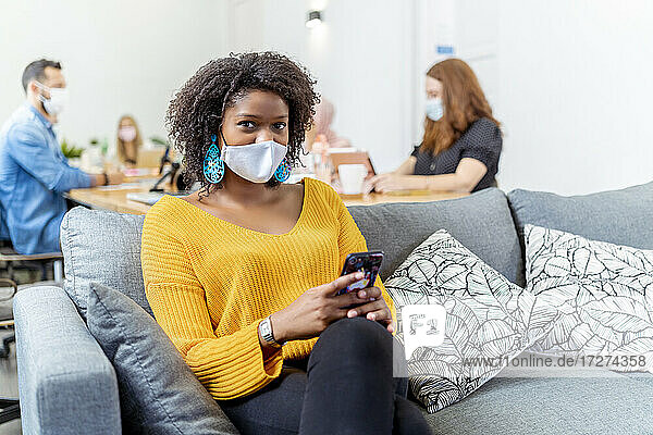 Mittlere erwachsene Frau mit Gesichtsmaske  die ein Mobiltelefon benutzt  während sie mit einem Mitarbeiter im Hintergrund im Büro sitzt