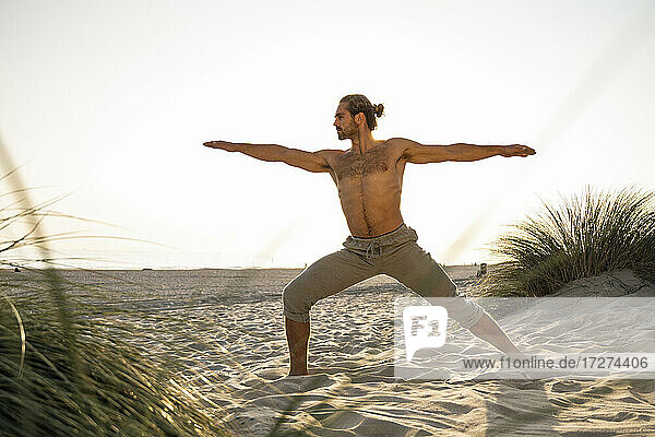 Shirtless man practicing warrior 2 position yoga at beach against clear sky during sunset