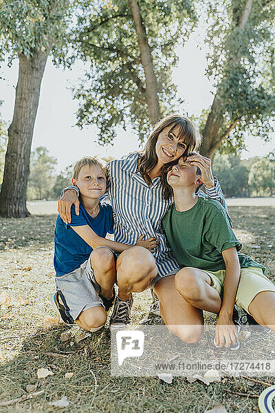 Happy mother embracing sons while kneeling in public park on sunny day