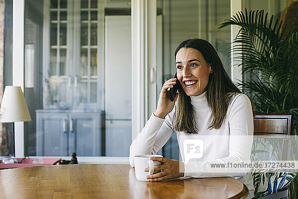 Junge Frau trinkt Kaffee  während sie zu Hause mit dem Handy telefoniert