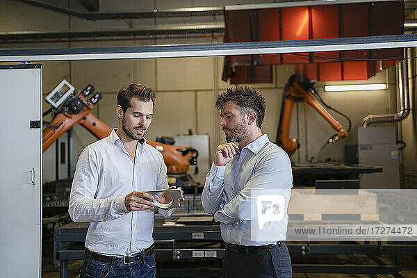 Businessman with metal object discussing with colleague while standing against robotic machine at factory