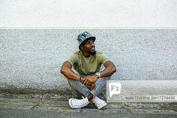 Nachdenklicher Mann mit Hut  der auf dem Fußweg gegen eine Mauer in der Stadt sitzt