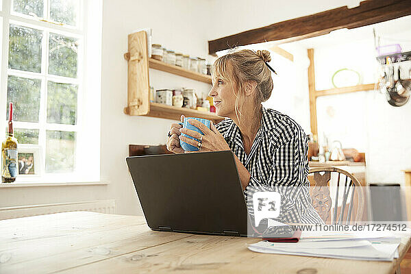 Ältere Frau  die wegschaut  während sie Kaffee trinkt und mit einem Laptop in der Küche zu Hause sitzt