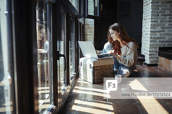 Junge Frau arbeitet am Laptop  während sie zu Hause auf dem Boden sitzt